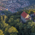 Drohnenbild der Wachenburg in Weinheim. Die Burg ist von der Sonne angestrahlt und umgeben von Wald. Im Hintergrund Blick auf Weinheim