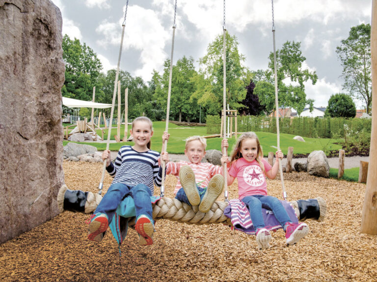 3 Kinder schaukeln auf einem natürlich angelegten Spielplatz und freuen sich.