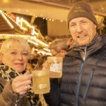 Ein Paar steht in einer Menschengruppe auf einem Weihnachtsmarkt und hat Glühweingläser in der Hand. Sie schauen in die Kamera