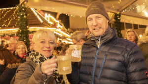 Ein Paar steht in einer Menschengruppe auf einem Weihnachtsmarkt und hat Glühweingläser in der Hand. Sie schauen in die Kamera