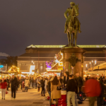 Gemütliche Abendstimmung auf dem Weihnachtsmarkt in Darmstadt. Menschen stehen zusammen und unterhalten sich. Blick auf die Reiterstatue von Großherzog Ludwig IV und das Hessische Landesmuseum auf dem Paradeplatz