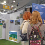 Messestand des Tourismus Service Bergstrasse e.V. An den Wänden hängen Bilder und Plakate, Broschüren liegen aus. Christiane Bartmann erklärt zwei Besucherinnen etwas.