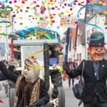 Verkleidete Fastnachtsfiguren laufen auf der Straße und winken. Jubelnde Zuschauer stehen am Straßenrand und feiern.