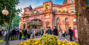 Zahlreiche Menschen stehe vor dem Kongresszentrum in Heidelberg. Die Sonne scheint und im Vordergrund blühen viele Blumen