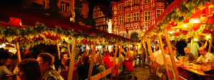 Zahlreiche Menschen sitzen gemütlich zusammen und trinken Wein. Alles ist festlich beleuchtet. Im Hintergrund abendlich beleuchtete Fachwerkhäuser