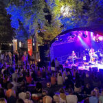 Zahlreiche Zuschauer sitzen vor einer Bühne auf der Musik gespielt wird. Schöne Atmosphäre in einem Park, der illuminiert ist