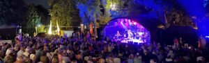 Zahlreiche Zuschauer sitzen vor einer Bühne auf der Musik gespielt wird. Schöne Atmosphäre in einem Park, der illuminiert ist