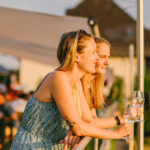 Zwei junge Frauen lehnen mit Weingläsern in den Händen entspannt über einem Geländer und schauen in die Ferne. Abendstimmung. Im Hintergrund sieht man verschwommen noch mehr Menschen.