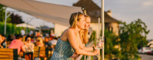 Zwei junge Frauen lehnen mit Weingläsern in den Händen entspannt über einem Geländer und schauen in die Ferne. Abendstimmung. Im Hintergrund sieht man verschwommen noch mehr Menschen.