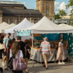 Auf dem Friedensplatz in Darmstadt stehen hübsche, einheitliche Zelte mit weißem Spitzdach. Darunter stellen Künstler Ihre Arbeiten aus. Zahlreiche Besucher sind vor Ort. Die Sonne scheint und die Menschen tragen sommerliche Kleidung