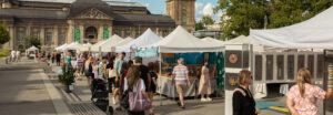 Auf dem Friedensplatz in Darmstadt stehen hübsche, einheitliche Zelte mit weißem Spitzdach. Darunter stellen Künstler Ihre Arbeiten aus. Zahlreiche Besucher sind vor Ort. Die Sonne scheint und die Menschen tragen sommerliche Kleidung
